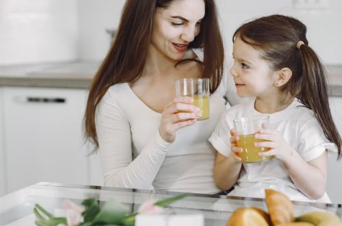 Happy mother and daughter in casual clothes drinking juice and looking at each other while having breakfast together in contemporary kitchen