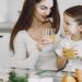 Happy mother and daughter in casual clothes drinking juice and looking at each other while having breakfast together in contemporary kitchen