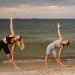 Two women exercising at a beach, performing yoga poses with the ocean in the background during sunset.